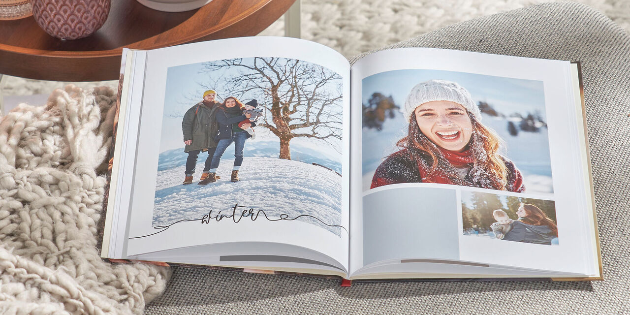 O CEWE FOTOCARTE deschisă se află pe o canapea gri. Fotocartea conține fotografiile a unei familii și cuvântul ”Iarnă”. În stânga este o masă cu lumănări și o plantă decorativă.